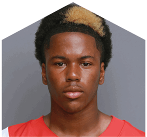 A person with short curly hair featuring a blonde patch is wearing a red sports jersey and looking directly at the camera against a plain gray background. Description generated by AI