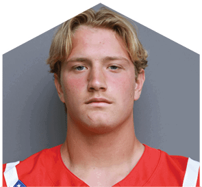 Person with short wavy blond hair wearing a red sports jersey with white accents against a plain gray background. Description generated by AI