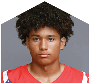 A person with curly hair wearing a red sports jersey looks directly at the camera against a plain gray background. Description generated by AI