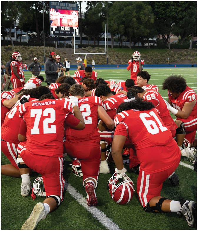 A group of people wearing red football uniforms are kneeling together on a field, some holding helmets, appearing to be in a moment of team unity or prayer. Description generated by AI