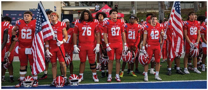 A group of people in red football uniforms stand in a line on a field holding American flags with helmets placed on the ground in front of them. Description generated by AI