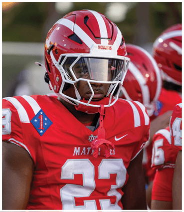 A person wearing a red football uniform with the number 23 and a helmet with a clear visor stands focused on the field. Description generated by AI