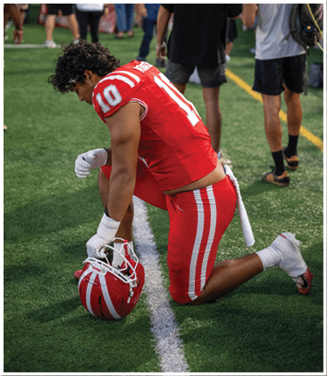 A person in a red sports jersey with the number 10 is bent over on a field while several people stand and walk in the background near a crowd. Description generated by AI