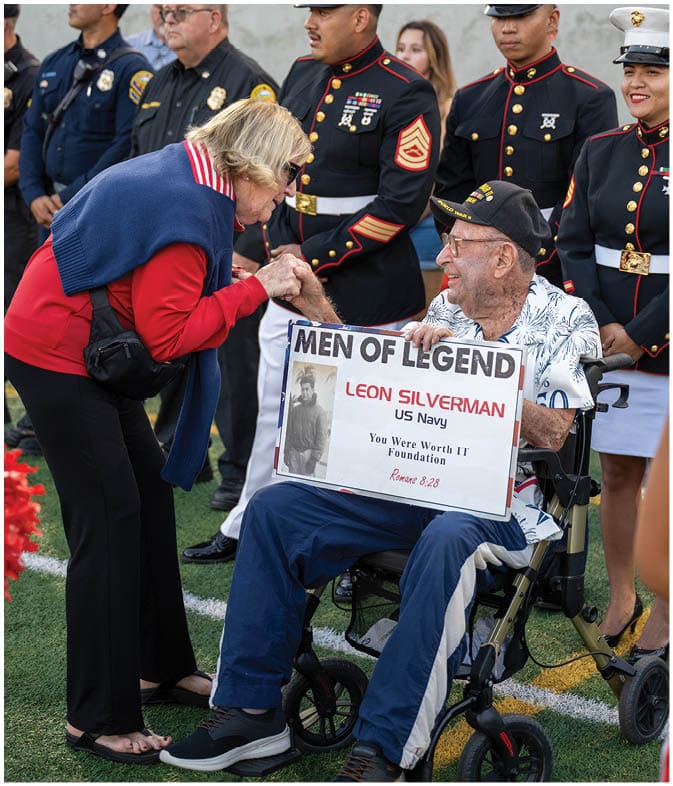 A group of people in military dress uniforms stand behind a senior wearing a cap and glasses who is holding a sign that reads “MEN OF LEGEND" while shaking hands with another person. Description generated by AI