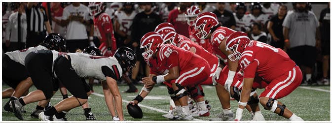Two opposing American football teams are lined up at the line of scrimmage with players in red uniforms facing players in white and black uniforms under stadium lights. Description generated by AI