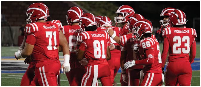A group of people wearing red football uniforms with helmets are gathered on a field, appearing to be in a team huddle or discussion. Description generated by AI