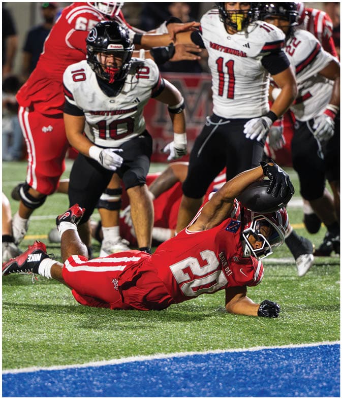 A person in a red football uniform is diving to catch a football near the end zone while several people in white and black football uniforms approach. Description generated by AI