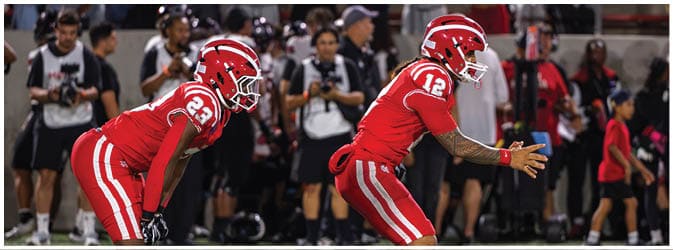Two people in red football uniforms and helmets are positioned on a field preparing for a play with a crowd and photographers in the background. Description generated by AI
