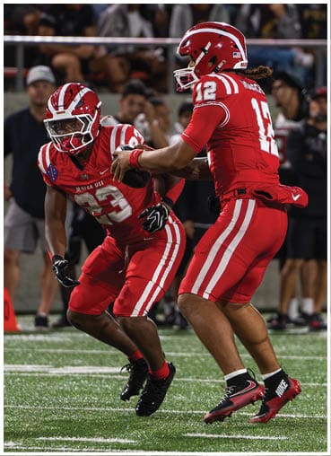 A person in a red and white football uniform is running on a field while holding a football and being handed off by another player in a similar uniform. Description generated by AI