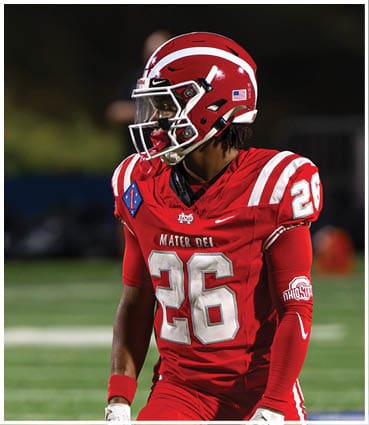 A person wearing a red football uniform with the number 26 and a red helmet stands on a field during a game. Description generated by AI