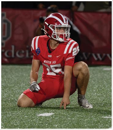 A person in a red football uniform and helmet is kneeling on a turf field with a blurred banner reading “Mater Dei" in the background. Description generated by AI