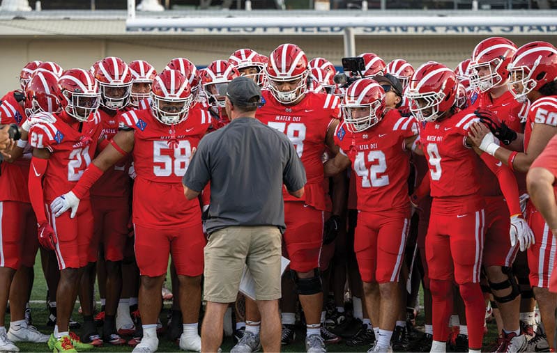 A group of people in red football uniforms and helmets stand closely together while a person in a gray shirt and shorts faces them, appearing to give instructions. Description generated by AI