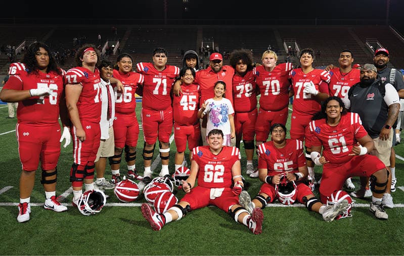 A group of people wearing red football uniforms and casual clothing pose together on a football field with helmets placed on the ground in front of them. Description generated by AI
