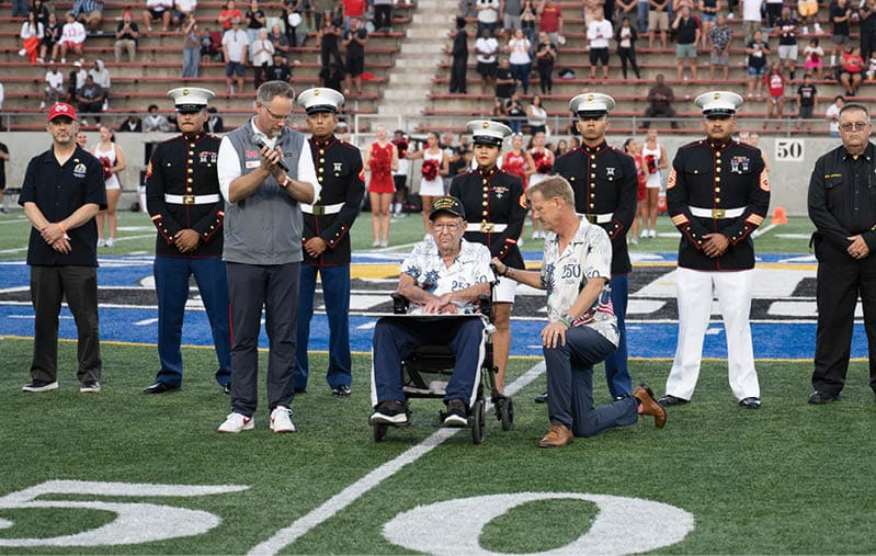 A group of people including uniformed military personnel, two adults kneeling, one adult standing with hands clasped, and a senior person in a wheelchair are gathered on a football field near the 50-yard line with spectators in the background. Description generated by AI