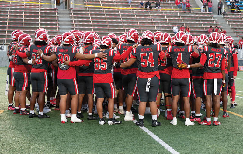 A group of people wearing red and black football uniforms and helmets are huddled together on a green field with empty bleachers in the background. Description generated by AI