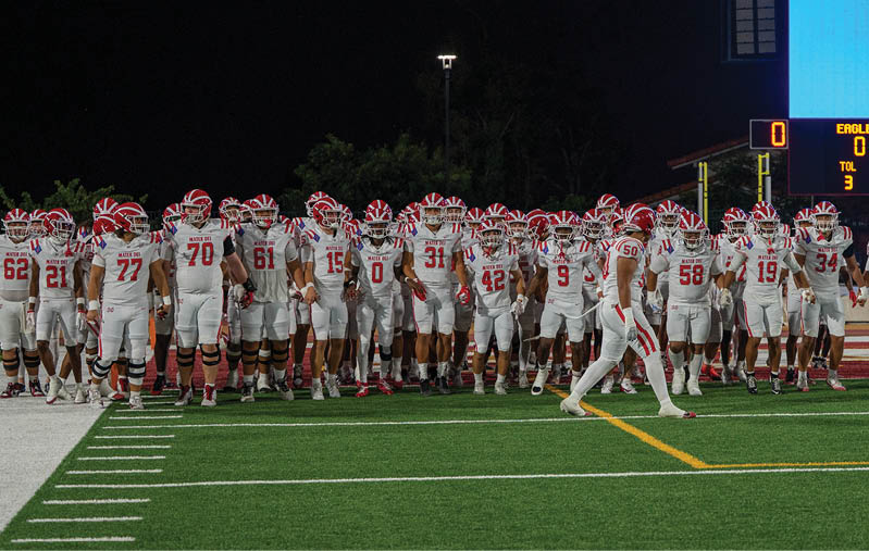 A large group of people wearing white football uniforms with red helmets are gathered on a football field at night near a scoreboard. Description generated by AI