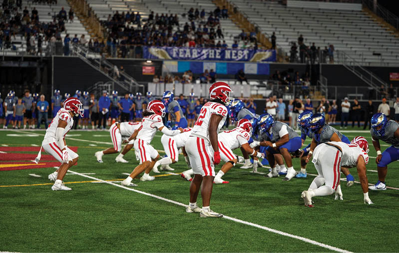 Players in red and white uniforms and players in blue uniforms are lined up on a football field preparing for a play during a game in a stadium with spectators in the stands. Description generated by AI