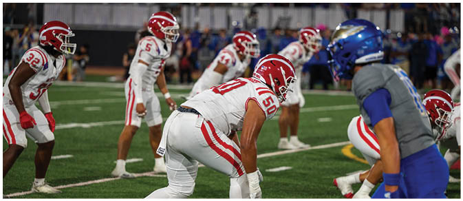 Several football players in white and red uniforms are positioned on a field facing a player in a blue uniform during a game. Description generated by AI
