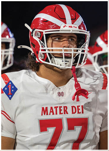 A person wearing a red and white football helmet and a white football jersey with red lettering that reads “MATER DEI" and the number 77 stands on a field with other players in the background. Description generated by AI