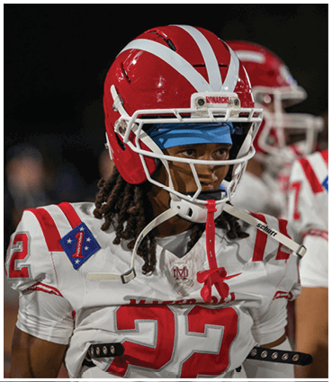 A person wearing a red and white football helmet and a white football jersey with the number 22 stands on a field with other players in the background. Description generated by AI