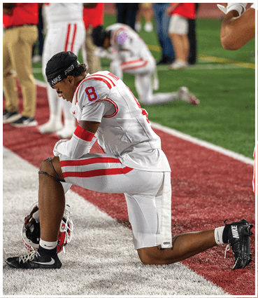 A person in a white and red football uniform is kneeling on one knee on a field with a helmet placed on the ground beside them. Description generated by AI