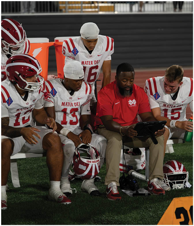 A group of football players in white and red uniforms sit on a bench with a coach in a red shirt showing them something on a tablet during a game. Description generated by AI