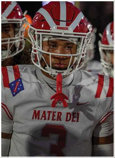 A person wearing a red and white football helmet with the word Monarchs on it and a white football jersey is looking intently to the side. Description generated by AI