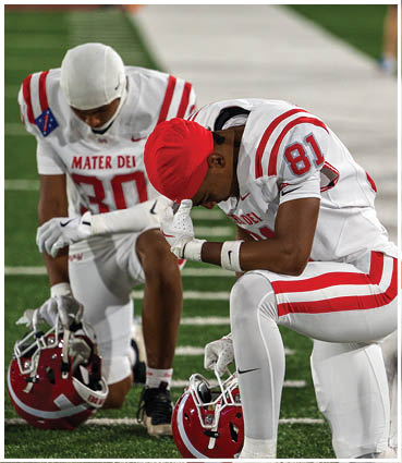 A person wearing a red cap and a white sports jersey with red stripes is bent forward, with another similarly dressed person partially visible in the background. Description generated by AI