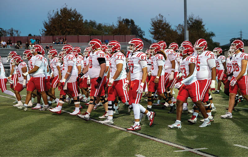 A group of football players in red and white uniforms and helmets are walking together on a grassy field during dusk. Description generated by AI