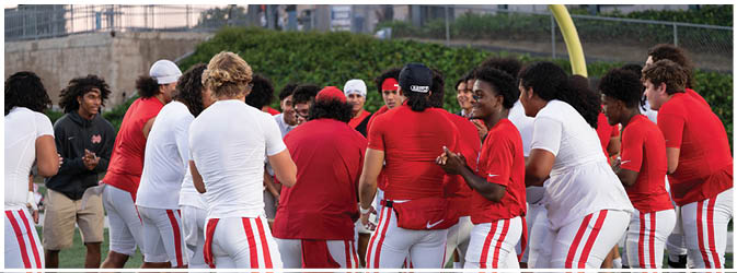 A group of people wearing red and white athletic clothing are gathered together on a sports field near a goalpost, appearing to be in discussion or preparing for an activity. Description generated by AI