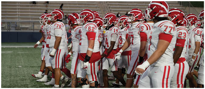 A group of people wearing white and red football uniforms and helmets are standing together on a field. Description generated by AI