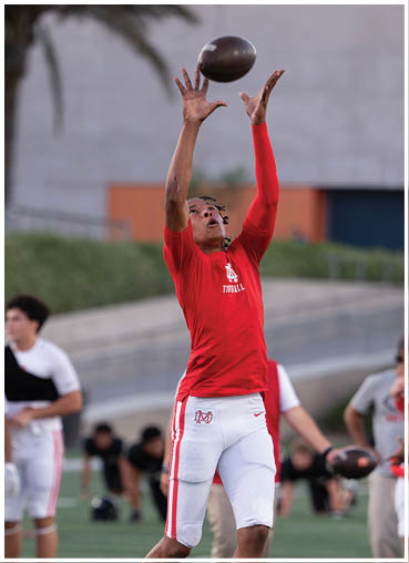 A person wearing a red long-sleeve shirt is reaching up with both hands to catch a football in an outdoor setting. Description generated by AI