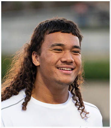 A person with curly hair styled in a mullet is smiling and wearing a white shirt outdoors. Description generated by AI