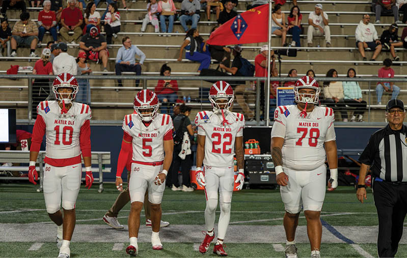 Four football players in white and red uniforms walk side by side on a field with spectators in the background. Description generated by AI