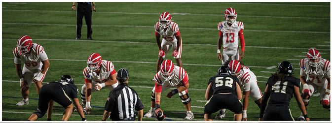 Two opposing American football teams are lined up at the line of scrimmage on a field under stadium lights with a referee standing nearby. Description generated by AI