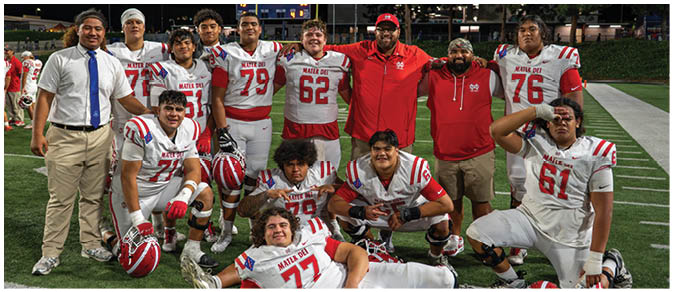 A group of adults in football uniforms and casual clothing pose together on a football field with some kneeling, standing, and one lying down. Description generated by AI