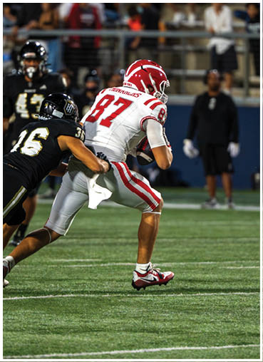 A person in a white and red football uniform is running while another person in a black football uniform is attempting to tackle them on a grassy field. Description generated by AI