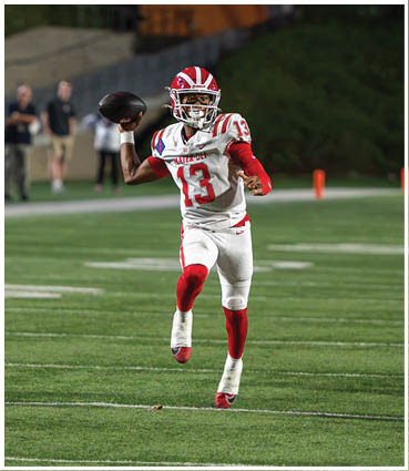 A person in a red and white football uniform is preparing to throw a football on a field while a referee and other players are nearby. Description generated by AI