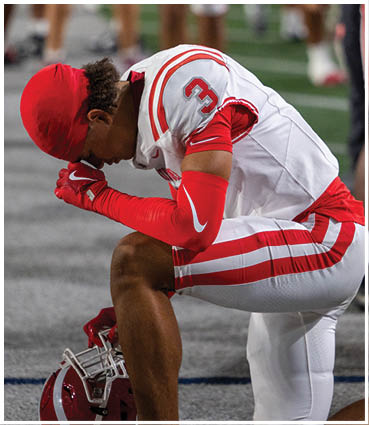 A person in a white and red football uniform with the number three is kneeling on one knee with their head bowed on a sports field. Description generated by AI