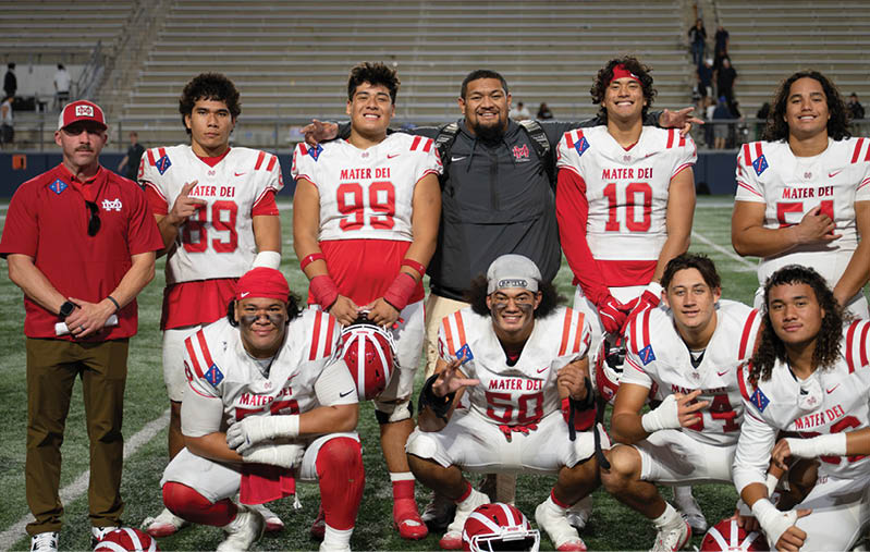 A group of people wearing football uniforms and one person in casual clothing pose together on a football field with empty bleachers in the background. Description generated by AI