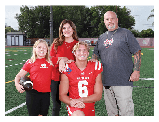 Four people smiling on a sports field with one person wearing a football uniform and the others wearing casual clothing with a sports team logo. Description generated by AI