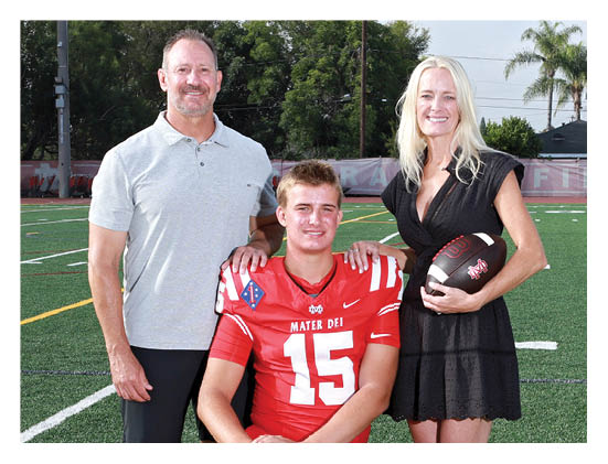 Three adults pose on a football field with one seated wearing a red football uniform and the other two standing on either side, one holding a football. Description generated by AI