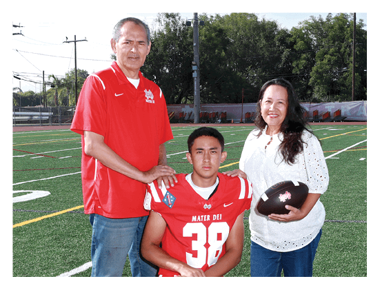 A person wearing a red football jersey with the number 38 is seated on a sports field while two adults stand beside them, one in a red polo shirt and the other holding a football and wearing a white blouse. Description generated by AI