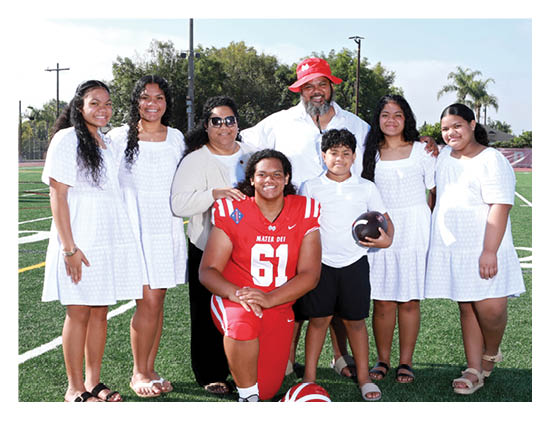 A group of eight people, including one person in a red football uniform kneeling with a helmet on the ground, pose together on a sports field with some wearing white dresses and others in casual clothing. Description generated by AI