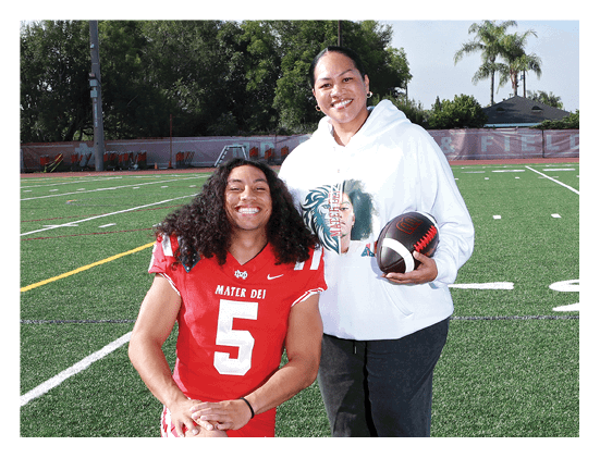 Two people are smiling on a football field, one wearing a red football jersey with the number five and the other wearing a white hoodie while holding a football. Description generated by AI