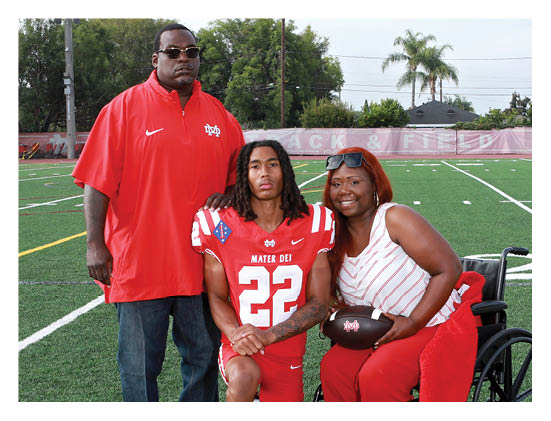 Three people pose together on a sports field with one person wearing a football uniform and the others standing and sitting close by, smiling. Description generated by AI