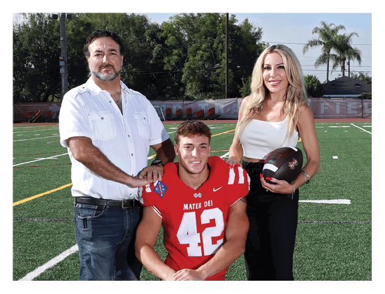 A person in a red football uniform kneels on a field between two standing adults, one wearing a white shirt and jeans and the other holding a football and dressed in a white top and black pants. Description generated by AI