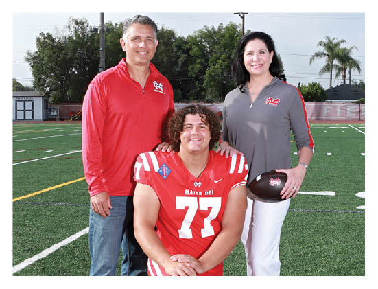 A person wearing a red football uniform with the number 77 is kneeling on a football field while two people stand behind them, one holding a football. Description generated by AI