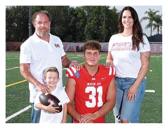 Four people stand together on a sports field with one person wearing a red football jersey and the others dressed casually, all smiling at the camera. Description generated by AI