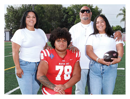 Four people stand on a football field with one person in a red football uniform sitting in front while the others stand behind wearing white shirts and jeans, with one holding a football. Description generated by AI